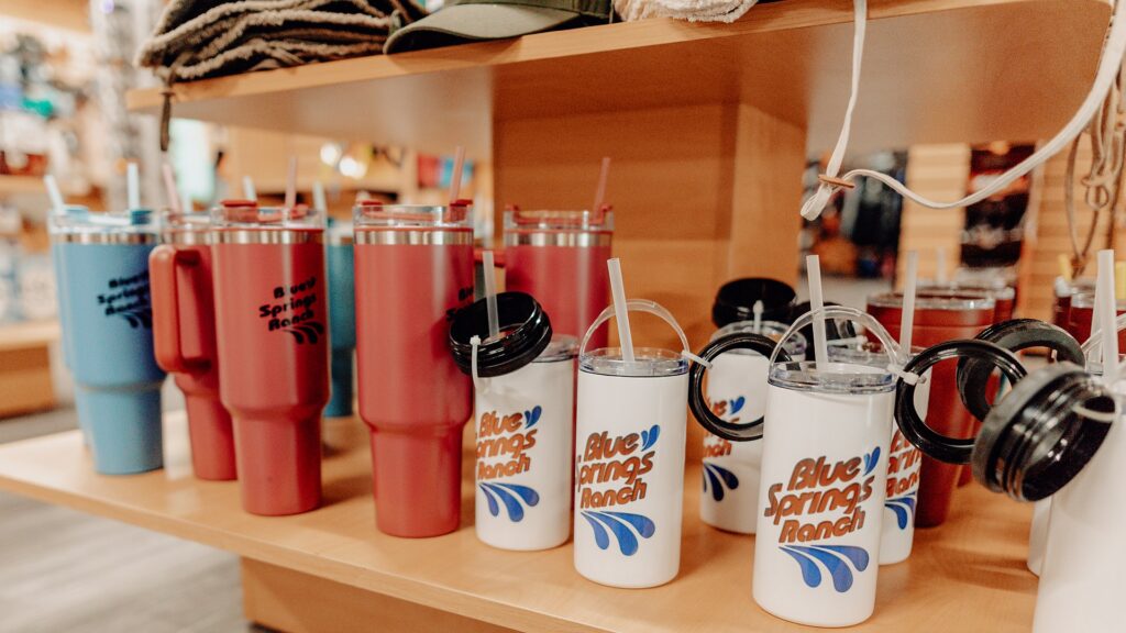 Blue Springs Ranch branded tumblers and water bottles on a shelf in the camp store.