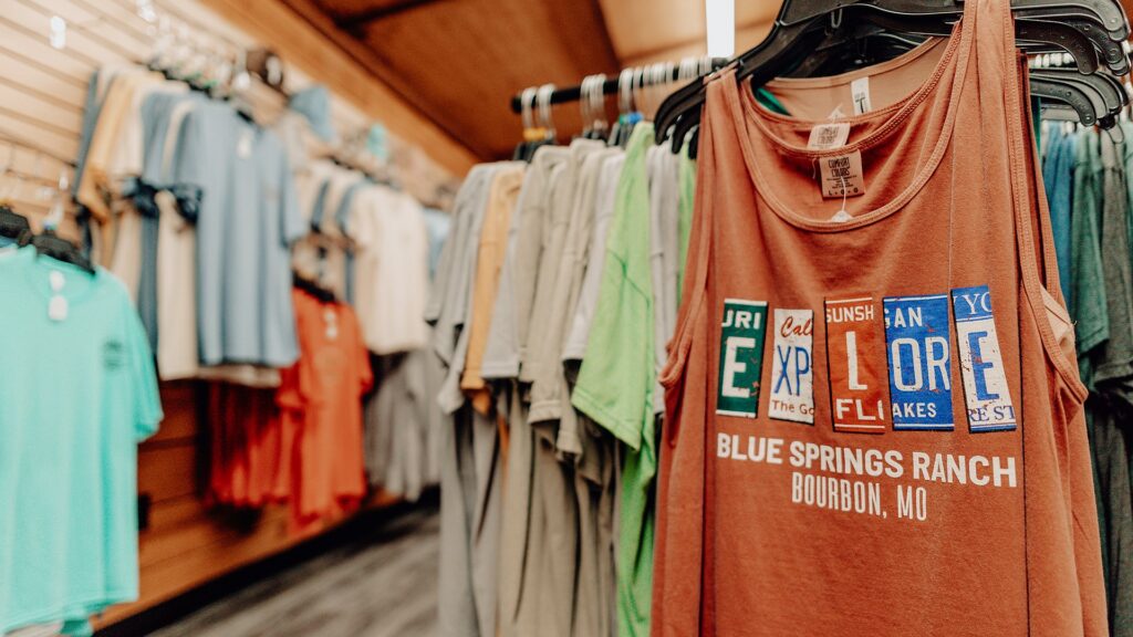 Racks of Blue Springs Ranch T-shirts in the camp store, an orange 'Explore' tee in front.