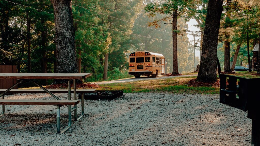 A campsite picnic table and fire ring with a shuttle bus passing on the road behind.
