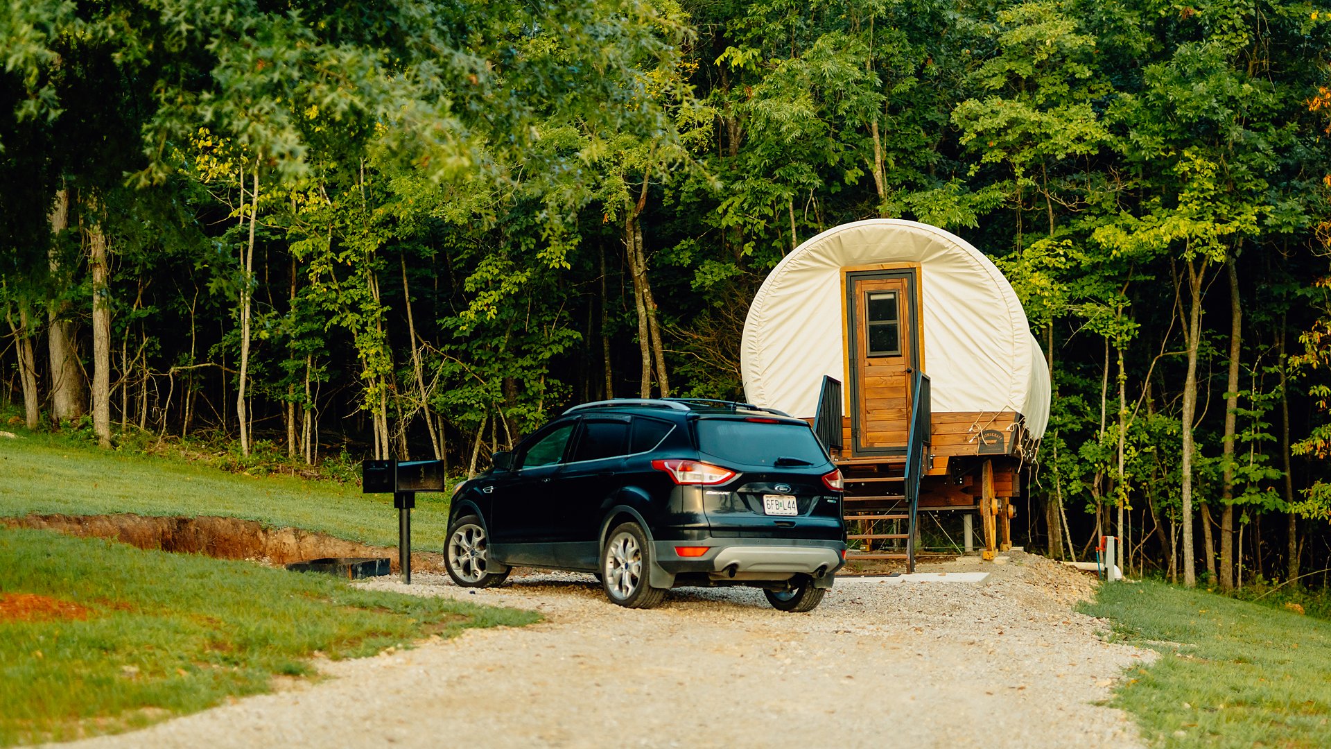 A canvas-topped Conestoga wagon in the woods with a car parked alongside.