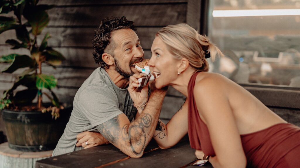 A couple sharing a s'more across a picnic table at dusk.