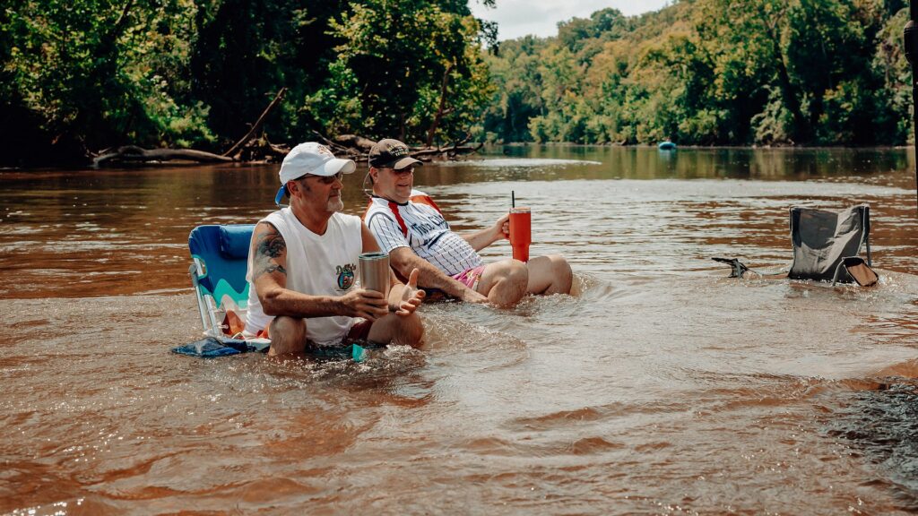 Two friends sitting in camp chairs in the shallow river with drinks.