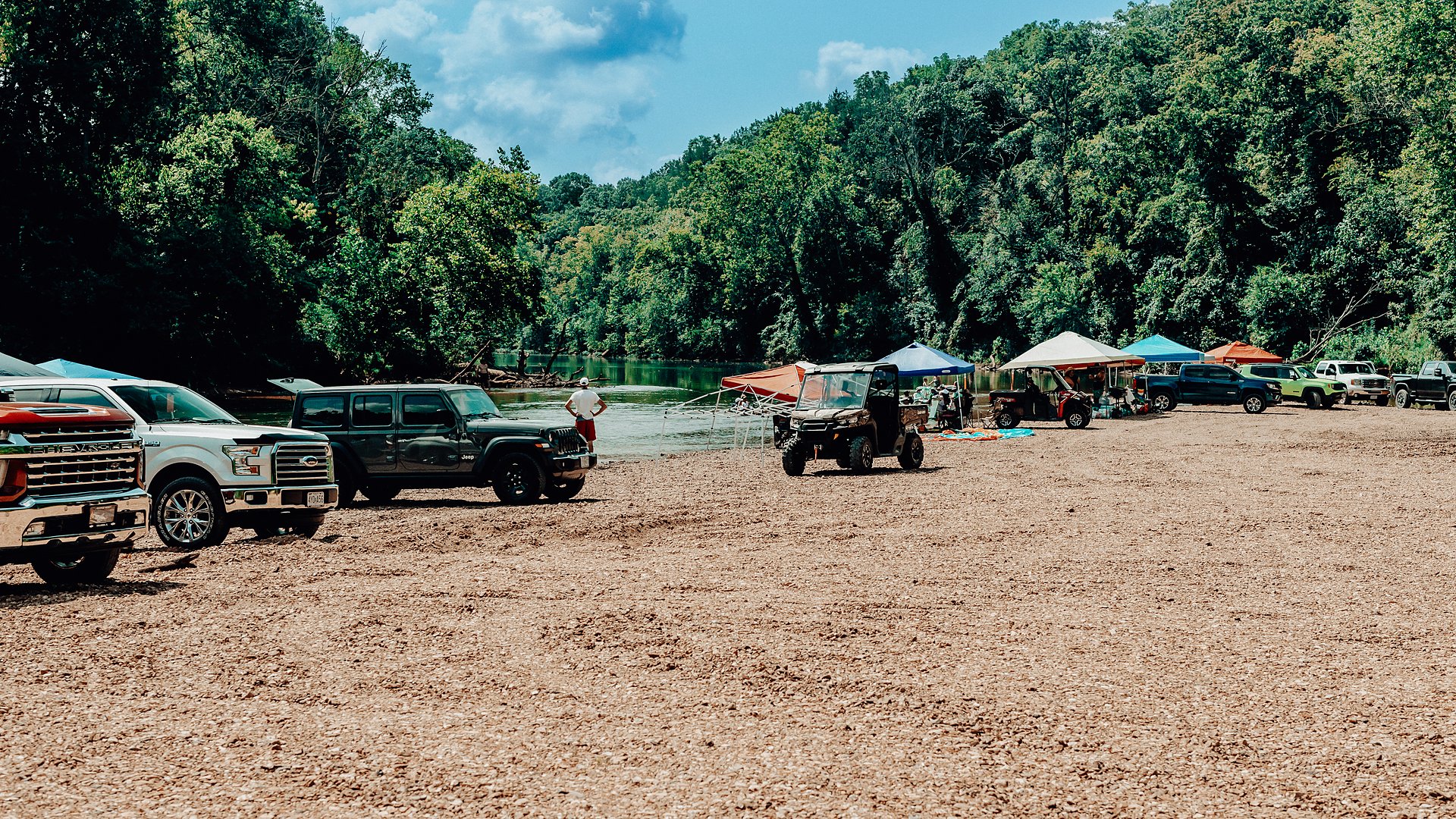 Vehicles and canopies spread along the gravel bar at the river's edge.