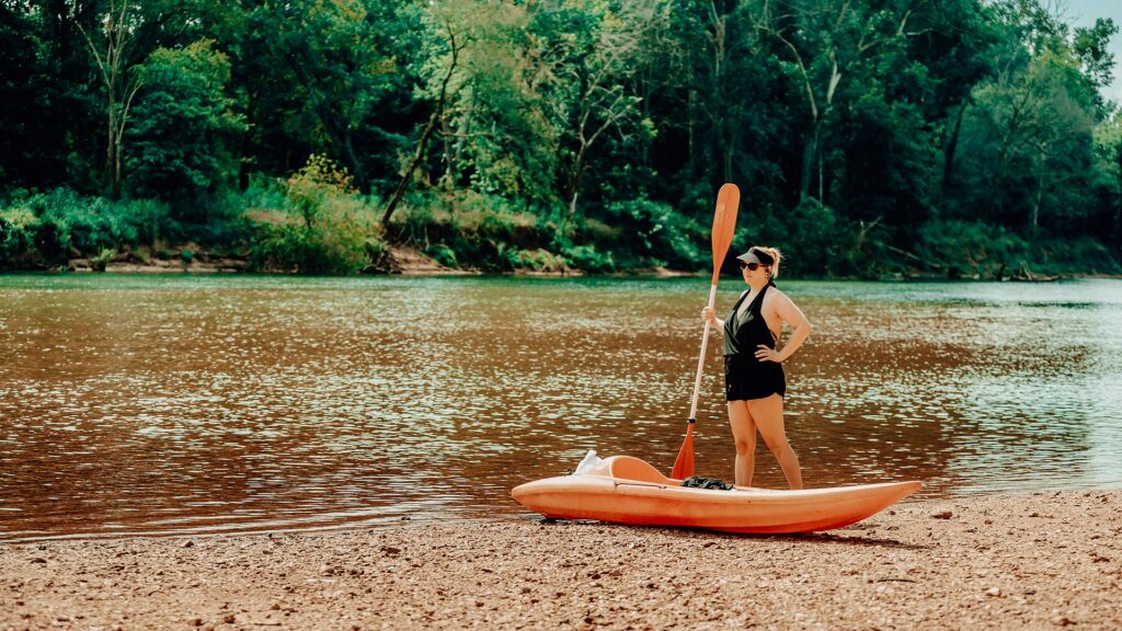 A guest standing with her paddle beside an orange kayak on the riverbank.