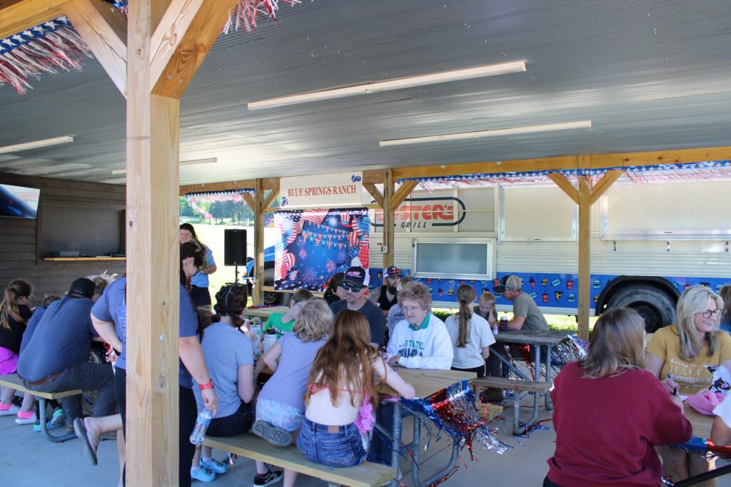 A crowded pavilion of families at picnic tables, with a Blue Springs Ranch sign and the grill bus behind.