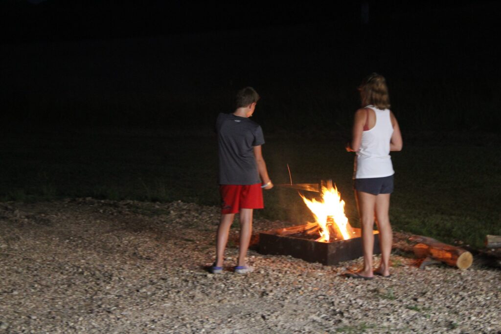 Two guests standing beside a blazing fire pit on the gravel at night.