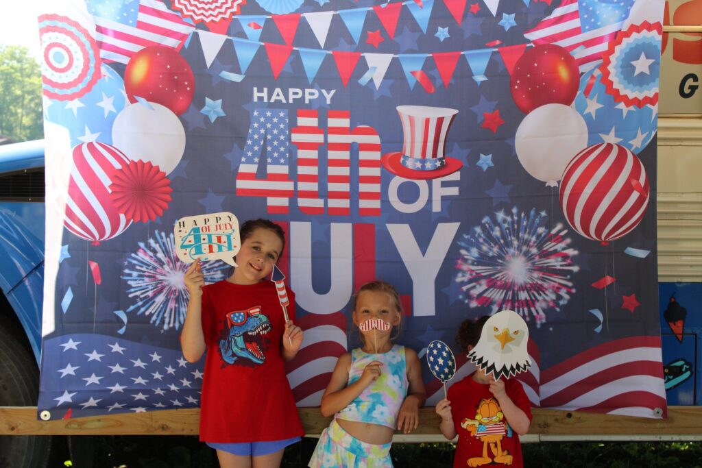 Three children holding party props in front of a Happy 4th of July photo backdrop.