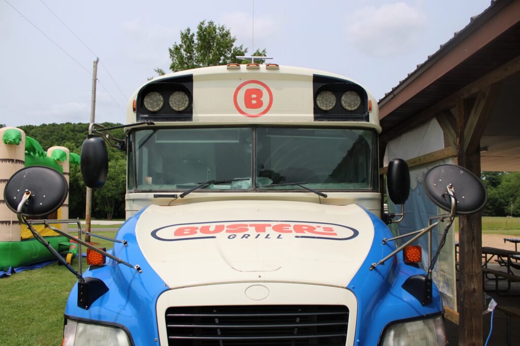 Front of the Buster's Grill bus parked beside a pavilion, with a bounce house behind.