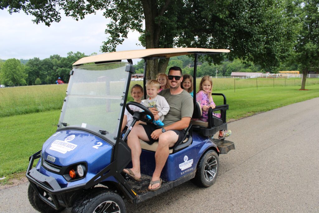 A man and several children smiling from a blue golf cart on a paved road.