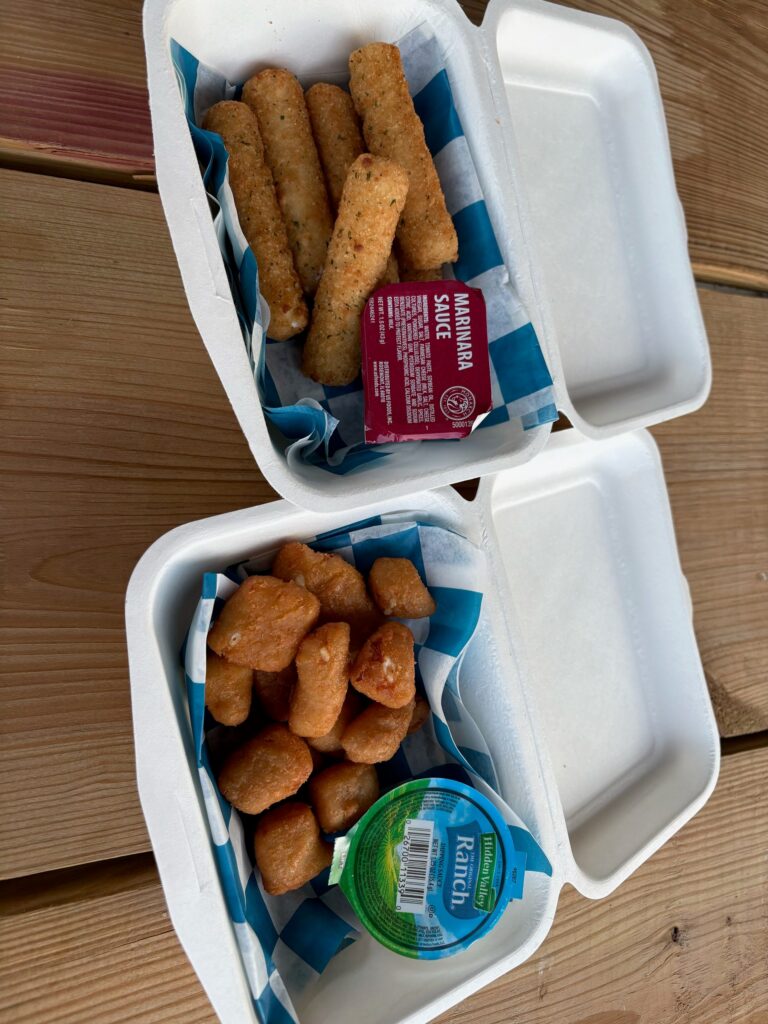 Two takeout boxes on a picnic table, one of mozzarella sticks and one of cheese curds.