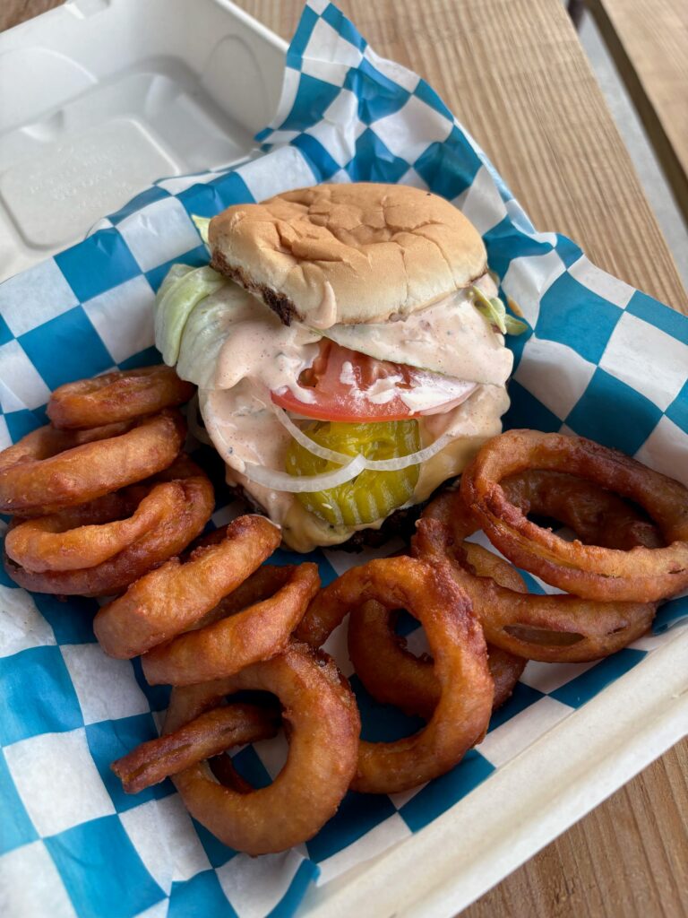 A smashburger with lettuce, tomato and pickles served with onion rings in a takeout box.