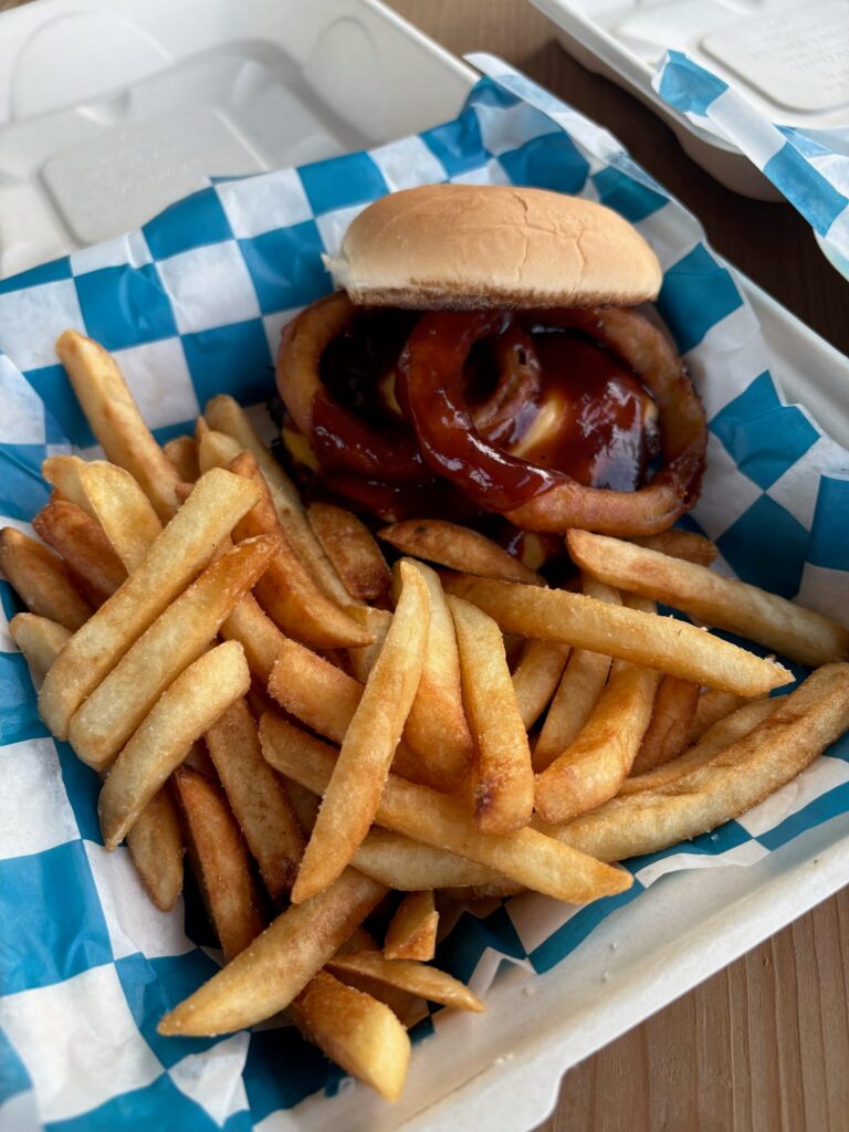 A western burger topped with onion rings and barbecue sauce beside a pile of fries.