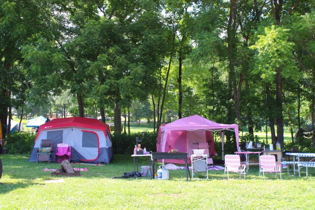 A red cabin tent and a pink canopy with picnic tables and folding chairs at a shaded tent site.