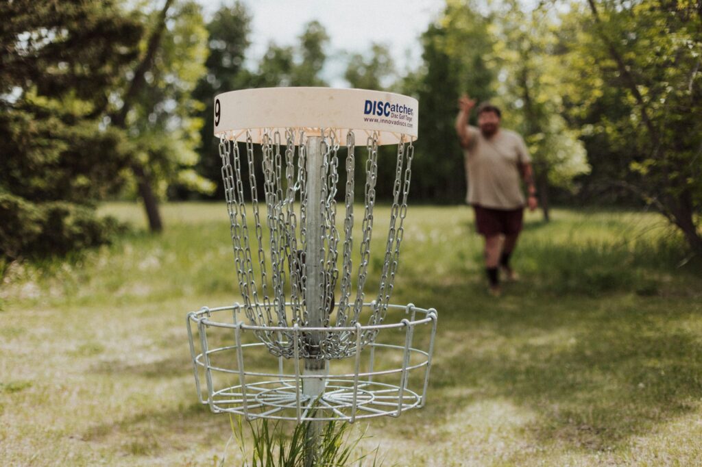 A disc golf basket on a grassy course, with a player throwing toward it from across the field.