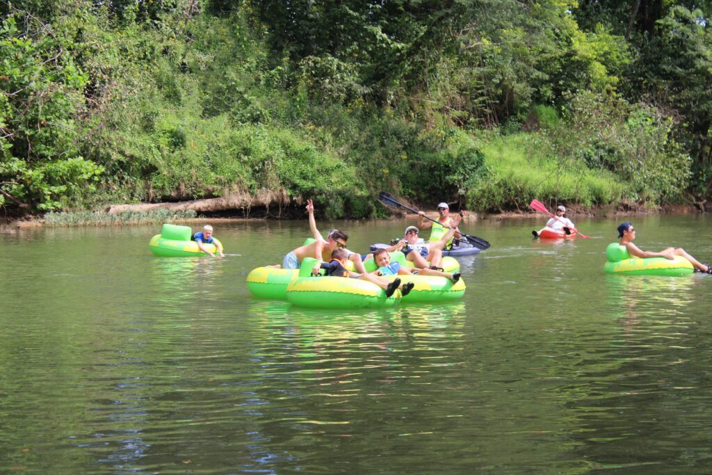 A group of guests floating together on green tubes, with kayakers paddling nearby.