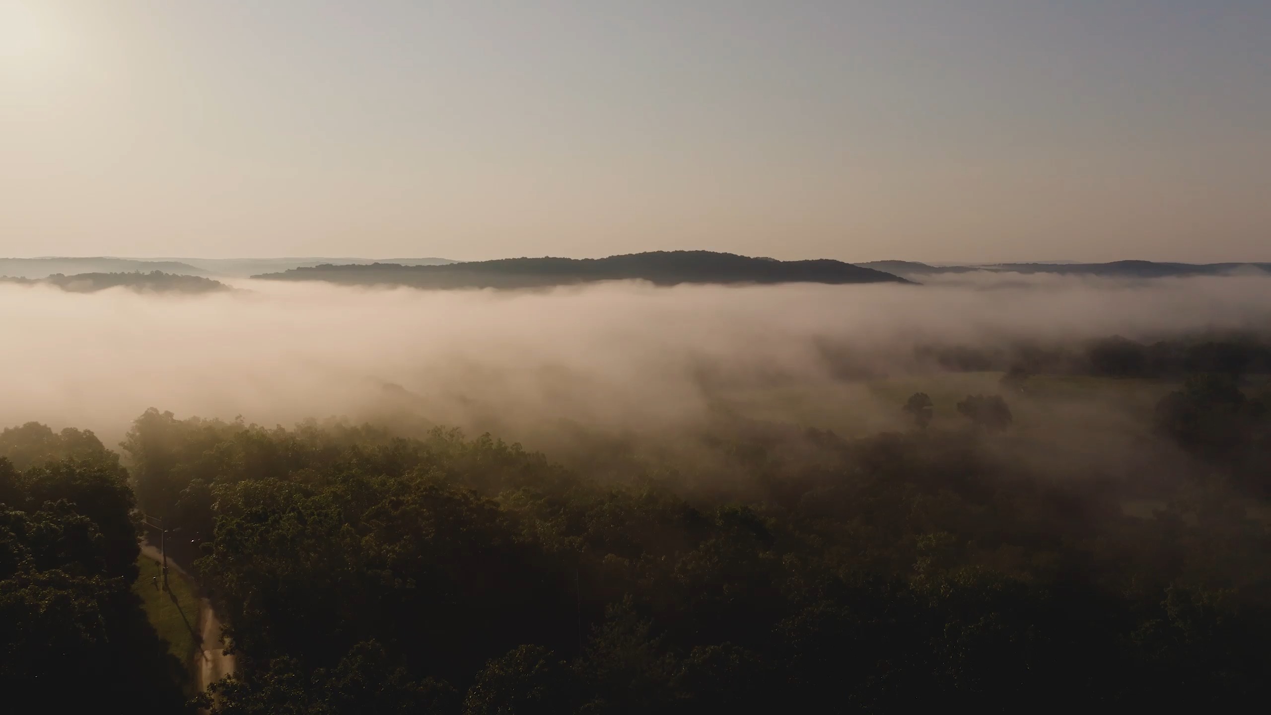 Aerial view of morning fog over the valley at dawn, with wooded hills beyond.