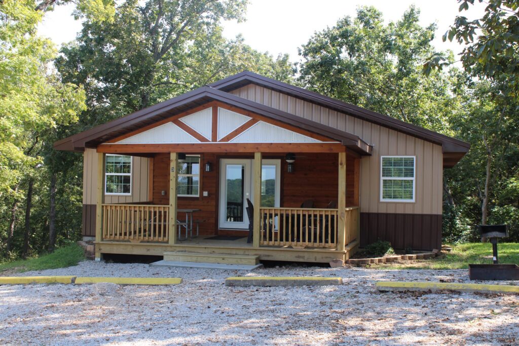 A tan and brown cabin with a covered front porch, a grill and gravel parking, among trees.