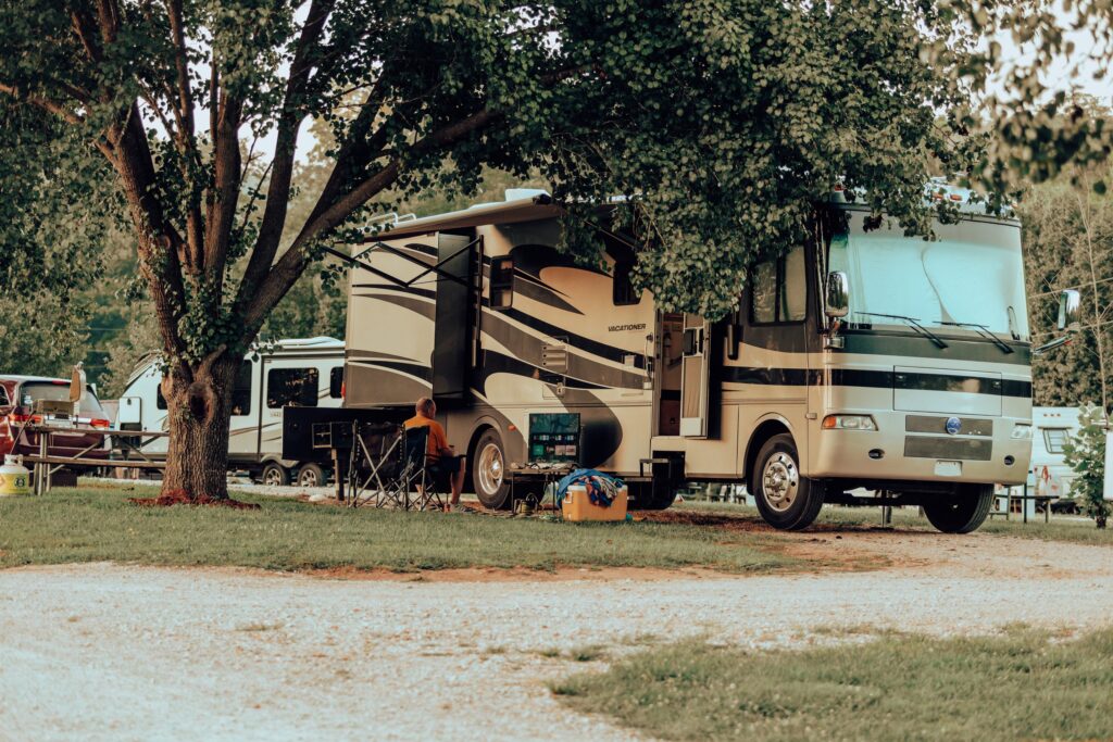 A motorhome parked under a shade tree at an RV site, a guest relaxing in a camp chair beside it